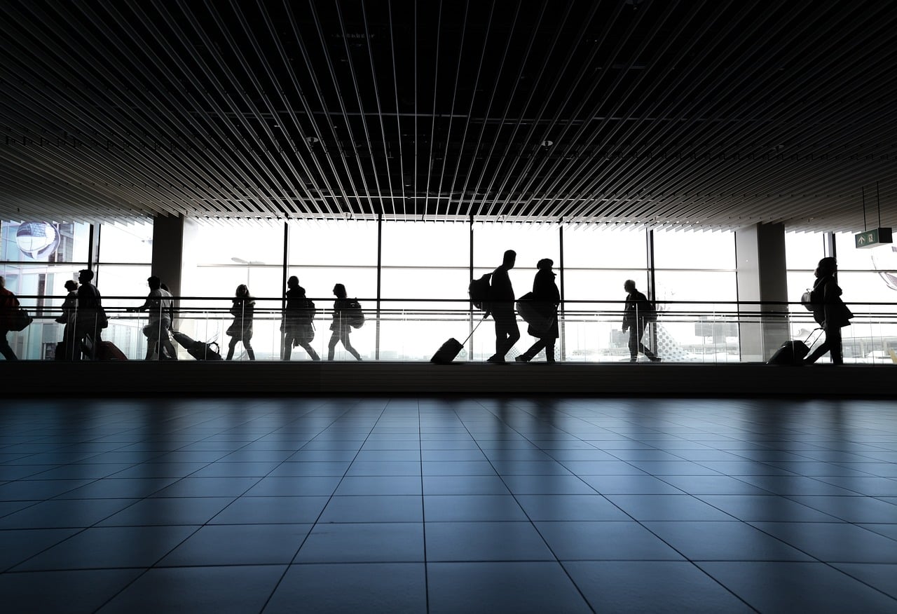 Travelers walking through airport terminal, silhouetted against window.